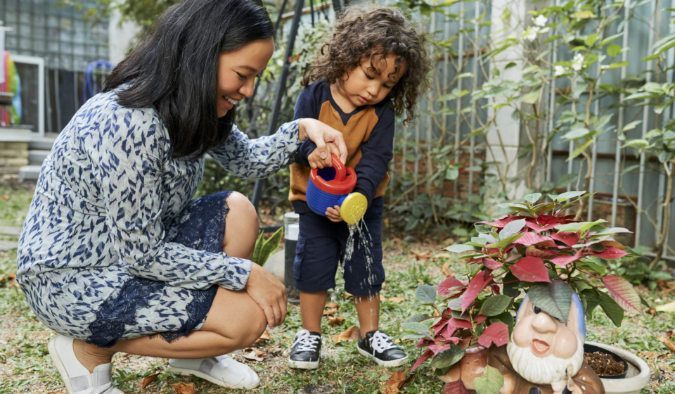 Smiling young mother teaching son how to water plants and flowers in backyard with watering can